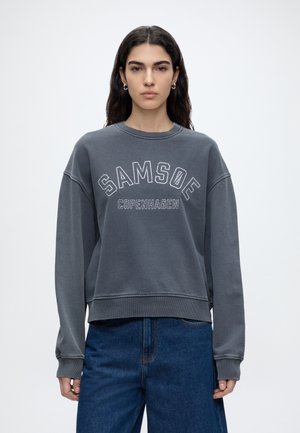 Giovane donna con capelli lunghi e scuri indossa una felpa grigia con la scritta "Samsoe Copenhagen" e jeans blu, in piedi davanti a uno sfondo chiaro e uniforme.