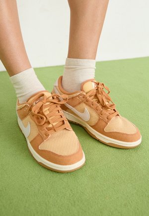 Baskets de sport marron en mesh et cuir suédé, avec des accents blancs, des lacets plats, un bout rond et une semelle rembourrée, portées avec des chaussettes claires.