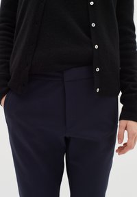 Černý cardigan na knoflíky, vrstvený přes tmavý top. Námořnické kalhoty hladké na dotek s předním skladem, s bočními kapsami.