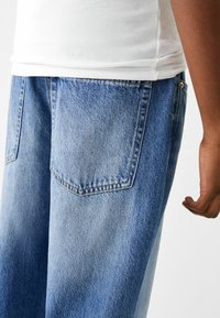 Jeans in denim azzurro chiaro con una vestibilità rilassata, dotati di due tasche posteriori e un leggero effetto sbiadito. Una canotta bianca a coste è visibile sopra i jeans.