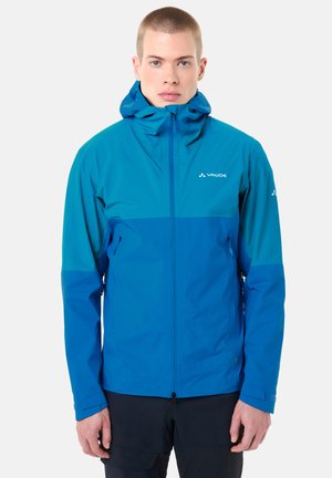 Giovane uomo che indossa una giacca impermeabile Vaude bicolore blu con cappuccio, in piedi e rivolto verso l'obiettivo su uno sfondo neutro.