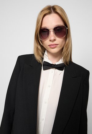 Donna con capelli biondi lisci che indossa occhiali da aviatore, blazer nero, camicia bianca abbottonata e papillon nero su sfondo grigio.