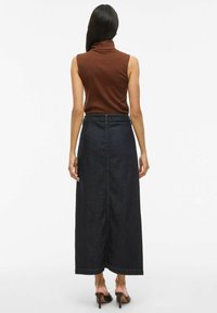 Femme de dos portant un haut à col roulé sans manches marron, une longue jupe en denim foncé et des sandales à talons marron.