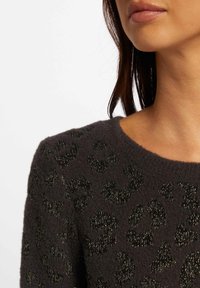 Femme portant un pull en maille foncée et duveteuse avec un imprimé léopard métallique noir, montrant le décolleté, l'épaule et une partie du visage avec de longs cheveux foncés.