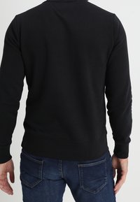 Sudadera negra con cuello redondo, puños y dobladillo acanalados, y una textura suave. Llevada con jeans azul oscuro que cuentan con bolsillos traseros.