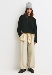 Felpa nera corta, pantaloni larghi beige, camicia gialla e bianca a righe annodata in vita, berretto grigio in maglia e scarpe scure.