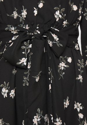 Tissu noir avec imprimé floral blanc et vert, orné d'un grand nœud noué à la taille sur un vêtement à manches longues.