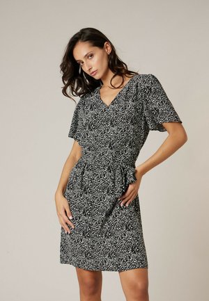 Vrouw met golvend donker haar, gekleed in een zwart-wit kort jurkje met korte mouwen, poseert met één hand op haar heup.