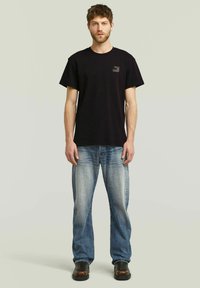 Camiseta de algodón negra con cuello redondo y mangas cortas. Llevada con jeans de mezclilla azul claro y zapatos negros. Detalle sutil del logo en la camiseta.