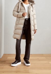 Abrigo acolchado beige con capucha, textura acolchada, cremallera frontal y detalles con cordón. Combina con vaqueros oscuros y zapatos negros de plataforma.