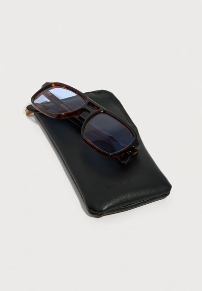 Τετράγωνα γυαλιά ηλίου σε χρώμα χελωνόσκαφου με μπλε φακούς, τοποθετημένα πάνω σε μαύρη θήκη από δέρμα γυαλιών, σε λευκό φόντο.