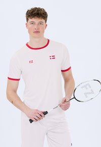 Nuori mies valkoisissa urheiluvaatteissa punaisilla yksityiskohdilla pitää badmintontikkua, päällään t-paita, jossa on Tanskan lippu ja "FZ"-logo.