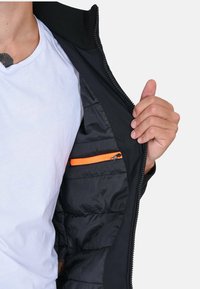 Giacca nera con cerniera, che mostra un interno trapuntato e una tasca con cerniera arancione. Materiale leggero, con colletto alto.