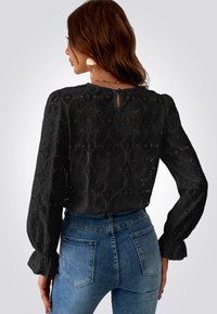 Zwarte blouse met lange mouwen, versierd met bloemachtige uitsnijdingen en een sleutelgatdetail aan de achterkant, gecombineerd met lichtblauwe high-waisted jeans.