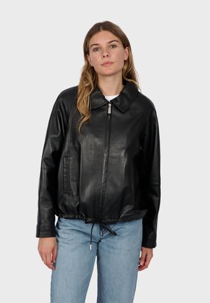 Junge Frau mit hellbraunem Haar trägt eine schwarze Lederjacke mit Reißverschluss und hellblaue Jeans vor einem einfarbigen hellen Hintergrund.