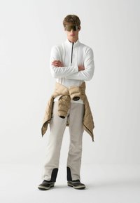 Sportna bela zip-up jakna, bež puffer jakna zavezana okoli pasu, svetlo obarvane hlače z temnimi akcenti in črne zimske čevlje.