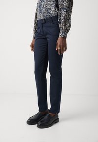 Pantalons navy avec une coupe ajustée, texture lisse et poches latérales. Associés avec des mocassins noirs, mettant en avant un design élégant et formel.
