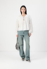 Weißes, strukturiertes Blusentop mit gebundenem Ausschnitt, kombiniert mit hellblauen, lässigen Jeans. Das Model hält eine silberne Handtasche und trägt metallische Schuhe.