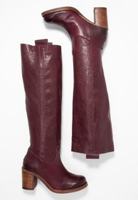 Knähöga burgundy läderstövlar med staplade träklackar, med en slät textur och sömdekor längs sidorna.