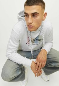 Grijze hoodie van zachte stof met een logo, trekkoordcapuchon en zwarte accenten; gecombineerd met lichtblauwe jeans en witte sneakers.