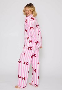 Conjunto de pijama a rayas verticales en rosa y blanco con grandes patrones de lazo en rojo. Parte superior de manga larga con bolsillo y pantalones de corte holgado. Tejido suave.