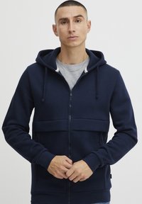 Felpa con zip di colore blu navy, dotata di tasca frontale, cappuccio con coulisse e polsini a coste. Caratterizzata da una texture liscia e un design minimalista.