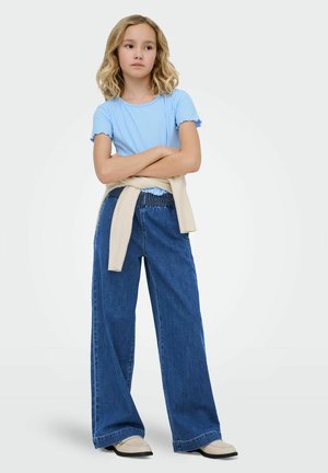 Plavokosa djevojka s valovitom kosom nosi svijetloplavu majicu, hlače širokih nogavica, bež džemper vezan oko struka i bež cipele, stoji s prekriženim rukama.