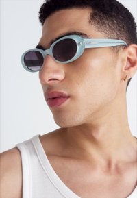 Akvino spalvos akiniai nuo saulės su apvaliais objektyvais, svelte dizainu ir kontrastingais tamsiai tonuotais objektyvais. Rėmelis turi mažą emblema.