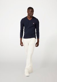 Marineblauw langemouwen V-hals shirt met een klein rood driehoekig logo, gecombineerd met witte broek en witte sneakers, gepresenteerd tegen een neutrale achtergrond.