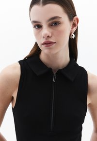 Haut sans manches noir avec une texture côtelée, doté d'une fermeture éclair et d'un col. Le modèle porte des boucles d'oreilles circulaires en argent.
