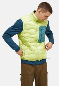 Hellgrüne gepolsterte Weste mit Wellenmuster, Reißverschlusstasche in Türkis, hohem Kragen und blauem Reißverschluss; getragen über einem blauen Langarmshirt.