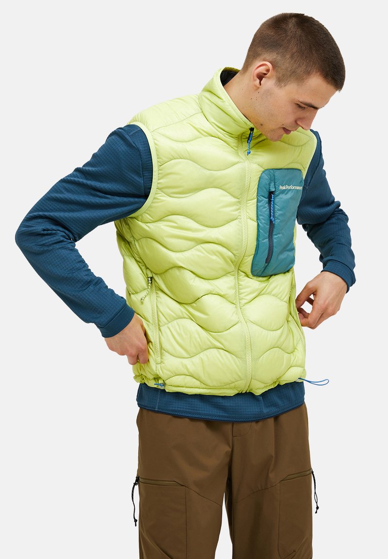 Hellgrüne gepolsterte Weste mit Wellenmuster, Reißverschlusstasche in Türkis, hohem Kragen und blauem Reißverschluss; getragen über einem blauen Langarmshirt.