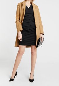 Manteau camel porté sur une robe noire ajustée, talons hauts et petite pochette noire. Le manteau présente un design structuré avec fermeture à boutons.