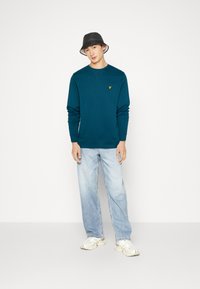 Felpa verde acqua con collo a girocollo, logo giallo sul petto; abbinata a jeans azzurri chiari e sneakers bianche; cappello a secchiello nero.
