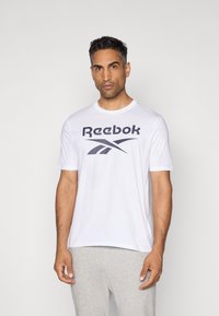 Weißes Reebok T-Shirt mit großem schwarzem Logo auf der Brust, kurzen Ärmeln und Rundhalsausschnitt. Getragen zu grauen Sweatpants.