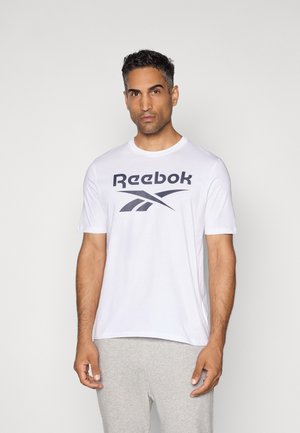 T-shirt blanc Reebok avec un grand logo noir sur la poitrine, manches courtes et col rond. Porté avec un pantalon de survêtement gris.