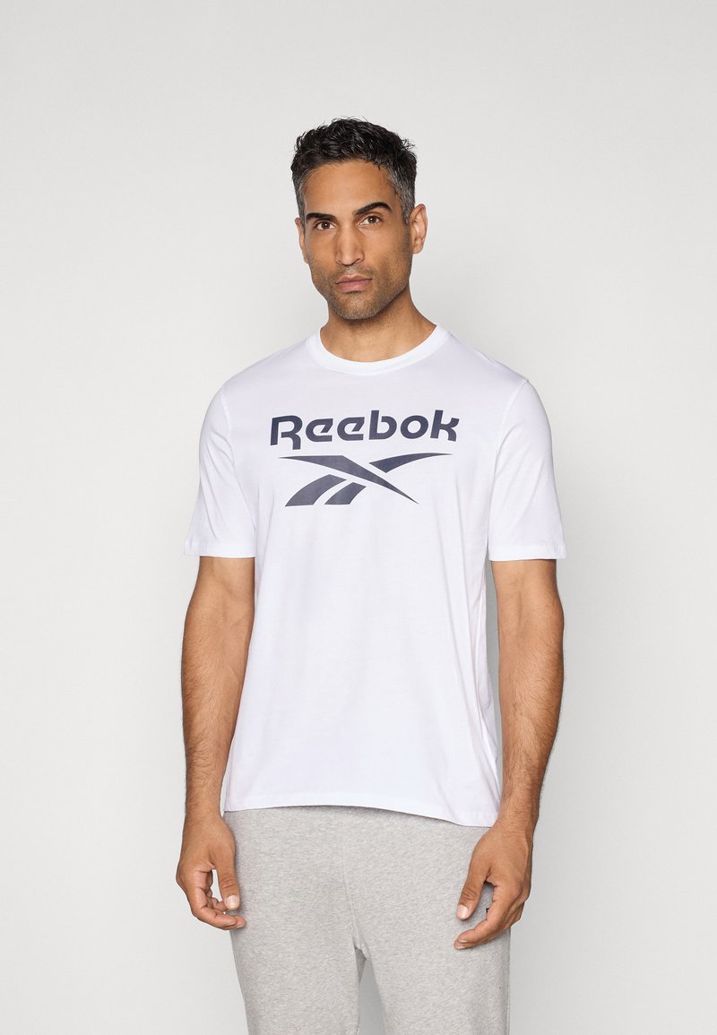Weißes Reebok T-Shirt mit großem schwarzem Logo auf der Brust, kurzen Ärmeln und Rundhalsausschnitt. Getragen zu grauen Sweatpants.