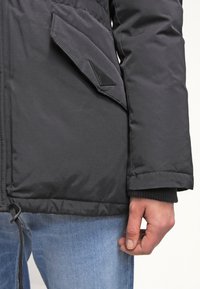 Black padded jacket featuring a zipper and a flap pocket, with ribbed cuffs and a drawstring hem. Fabric has a smooth texture.