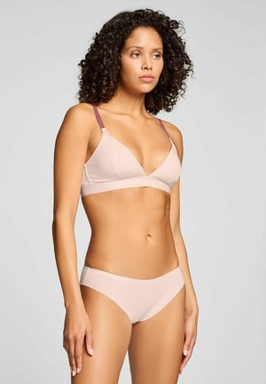Woman with curly hair wearing light pink bra with mauve straps and matching light pink briefs, standing against plain gray background.