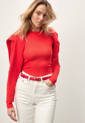 Punane pikkade varrukatega särk punase kampsuni all, valgete kõrge vöökohaga teksade ja neid värvi punase vööga, millel on sile tekstuur.