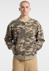Camo bomberjack in tinten van groen en bruin. Beschikt over een voorrits, geribde manchetten en zijzakken. Zacht, textuurstof.