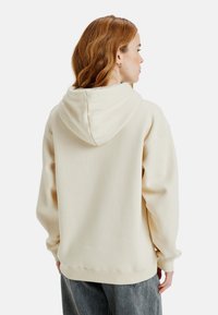 Crème-kleurige hoodie gemaakt van zacht materiaal, met een capuchon, een relaxte pasvorm en geribde manchetten. De achterkant toont een eenvoudig, minimalistisch ontwerp.