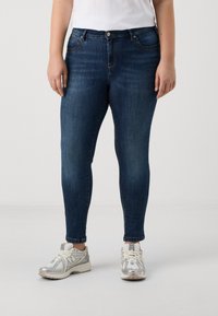 Skinny blue jeans made of denim with subtle fading, five-pocket design, front button closure, paired with gray and white sneakers.