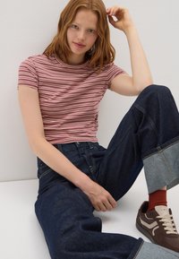 Jonge vrouw met rood haar in een gestreept T-shirt met korte mouwen, donkere opgerolde jeans, roestkleurige sokken en bruine sneakers, zittend tegen een witte muur.