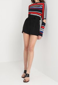 Pruhovaný top s dlouhým rukávem v červené, modré a bílé barvě; černé kraťasy s vysokým pasem a uvázaným páskem; černé sandály s otevřenou špičkou.