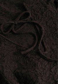 Tissu marron foncé avec une texture de dentelle florale en relief et un fin cordon ou ruban assorti, noué lâchement à la surface.