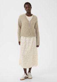Beiger Mohair-Strickjacke mit V-Ausschnitt, geripptem Saum und drei Knöpfen über einem weißen Maxikleid mit Blumenmuster, kombiniert mit weißen Sneakern.