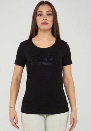 Tânără cu părul lung și drept, purtând un tricou negru cu mâneci scurte cu logo-ul EA7 Emporio Armani și pantaloni deschiși la culoare.