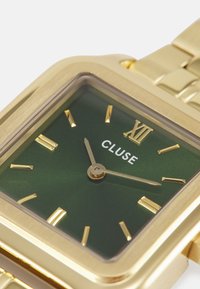 Goldfarbene rechteckige Uhr mit grünem Ziffernblatt, goldenen Stundenmarkierungen und dem prominenten Schriftzug "CLUSE". Metallarmband.