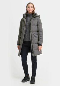 Grå polstret lang kåpe med hette, som har en teksturert strikkegenser under, sammen med mørke bukser og svarte sneakers.